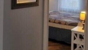 A narrow hallway leading to a bedroom with a bed, a bedside table with a lamp, and a ceiling light fixture.