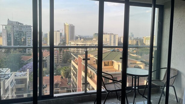 Un balcón con mesa y sillas, ventanas amplias con vista a la ciudad y piso de baldosas.