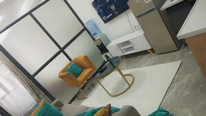 Una sala de estar moderna con un sofá, una mesa redonda y una silla. Hay un dispensador de agua y un televisor en la pared.