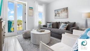 A modern living room with a grey sofa, a round coffee table, and a view through a glass door to the outside. - On Beach Time - New Construction - Deeded Beach Access / 3 Pools (Santa Rosa Beach)