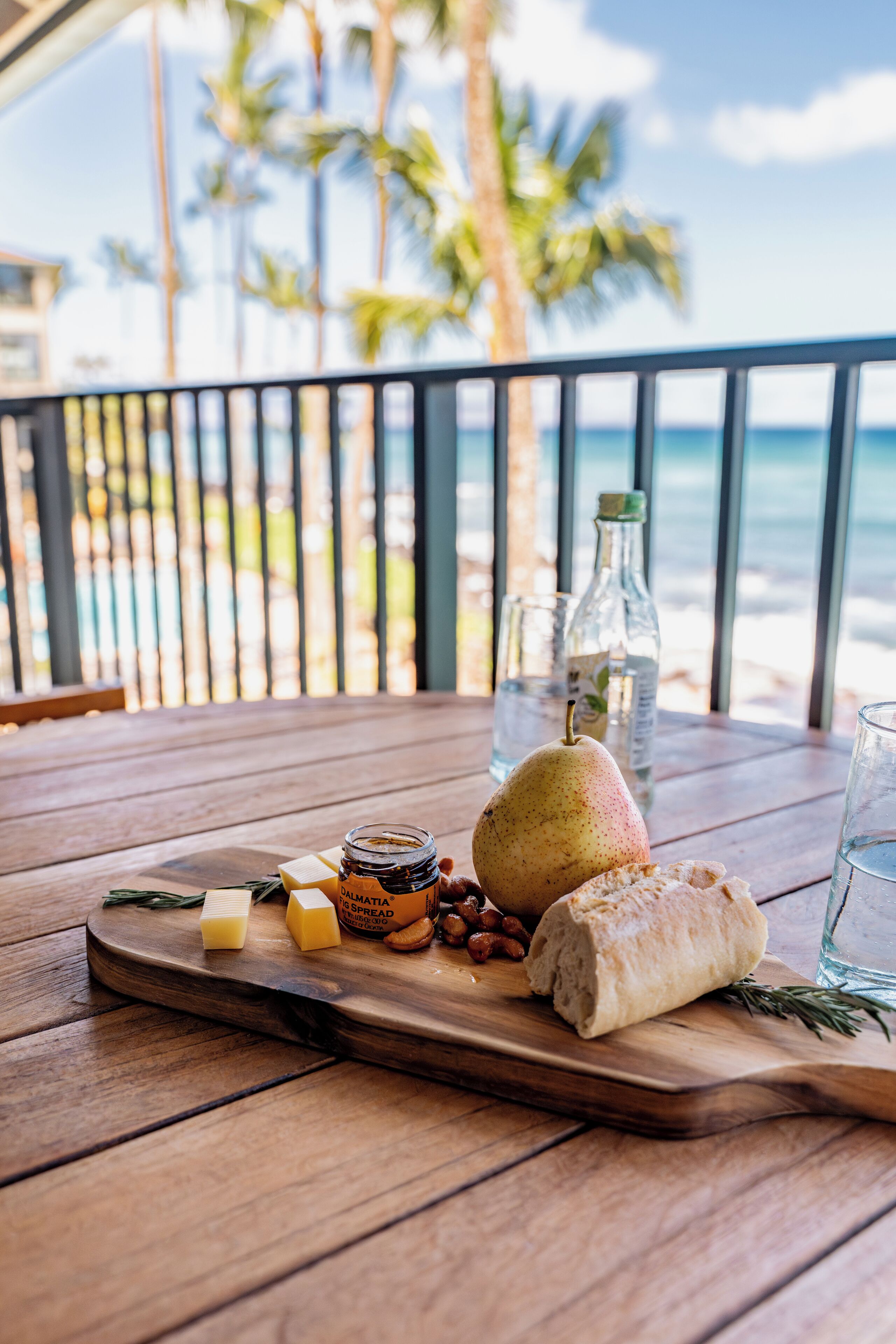 A wooden table with a cheese board, a pear, nuts, and a bottle of olive oil on a balcony overlooking a beach with palm trees.