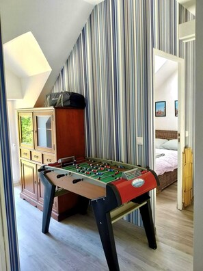 Una habitación con mesa de fútbolín, un armario con cajones, una cama con edredón blanco y una pared a rayas.