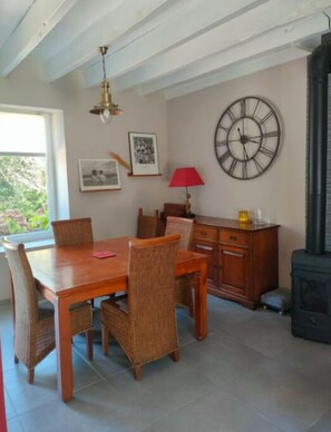 A dining area with a wooden table and wicker chairs, a lamp, and a large wall clock.