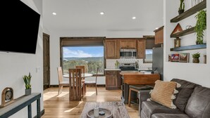 Ein modernes Wohnzimmer mit Sofa, Couchtisch und Essbereich mit Stühlen. Im Hintergrund ist eine Küche mit Holzausstattung und Blick ins Freie zu sehen.