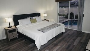 A bedroom with a bed, two bedside tables with lamps, a wooden nightstand, a gray throw blanket, and a sliding glass door leading to a pool area.