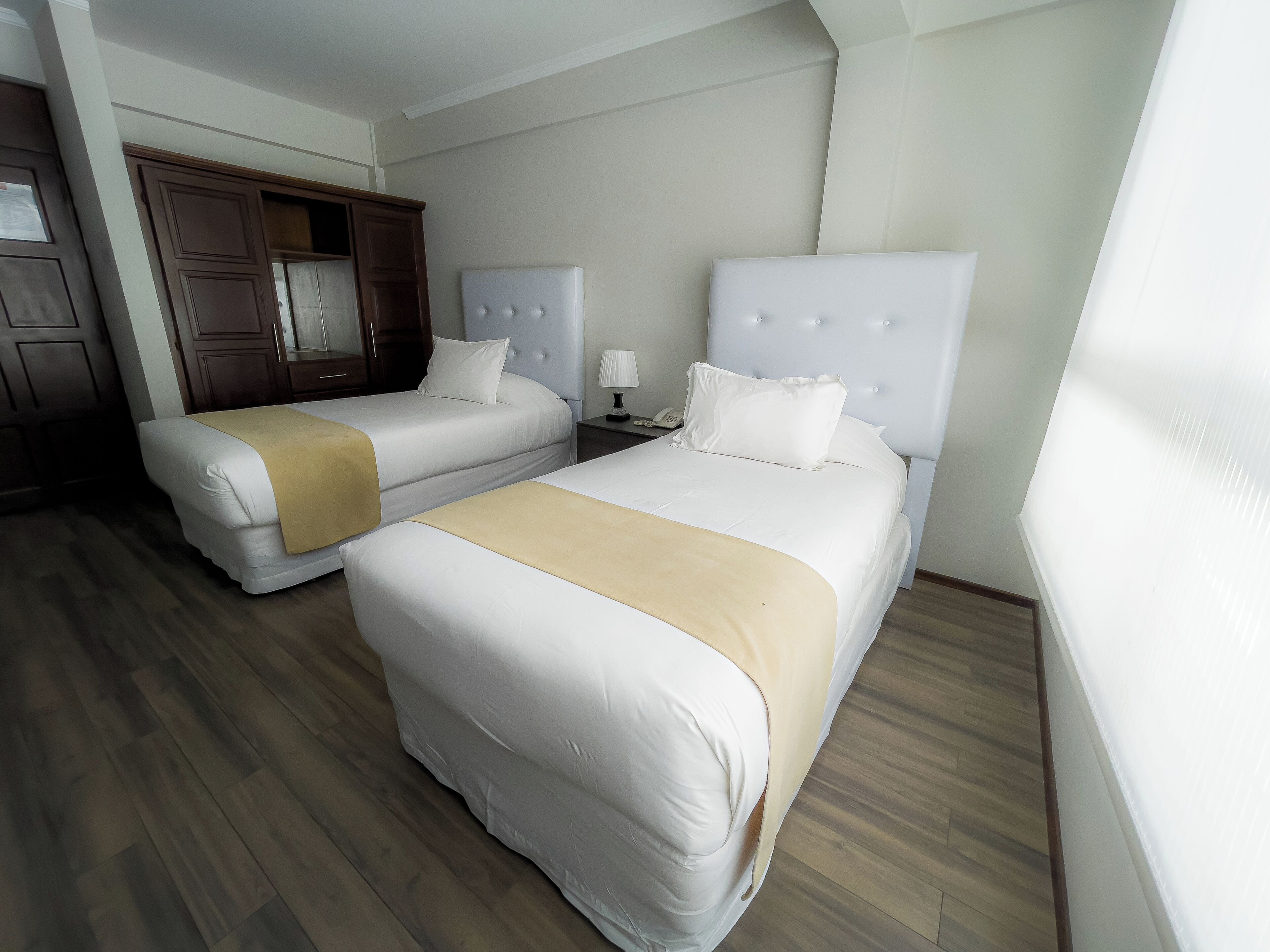 Habitación de hotel con dos camas, suelo de madera, un armario y una mesita de noche con lámpara.