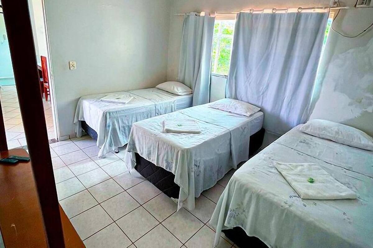 Une chambre avec trois lits simples, un sol carrelé et une fenêtre avec des rideaux.