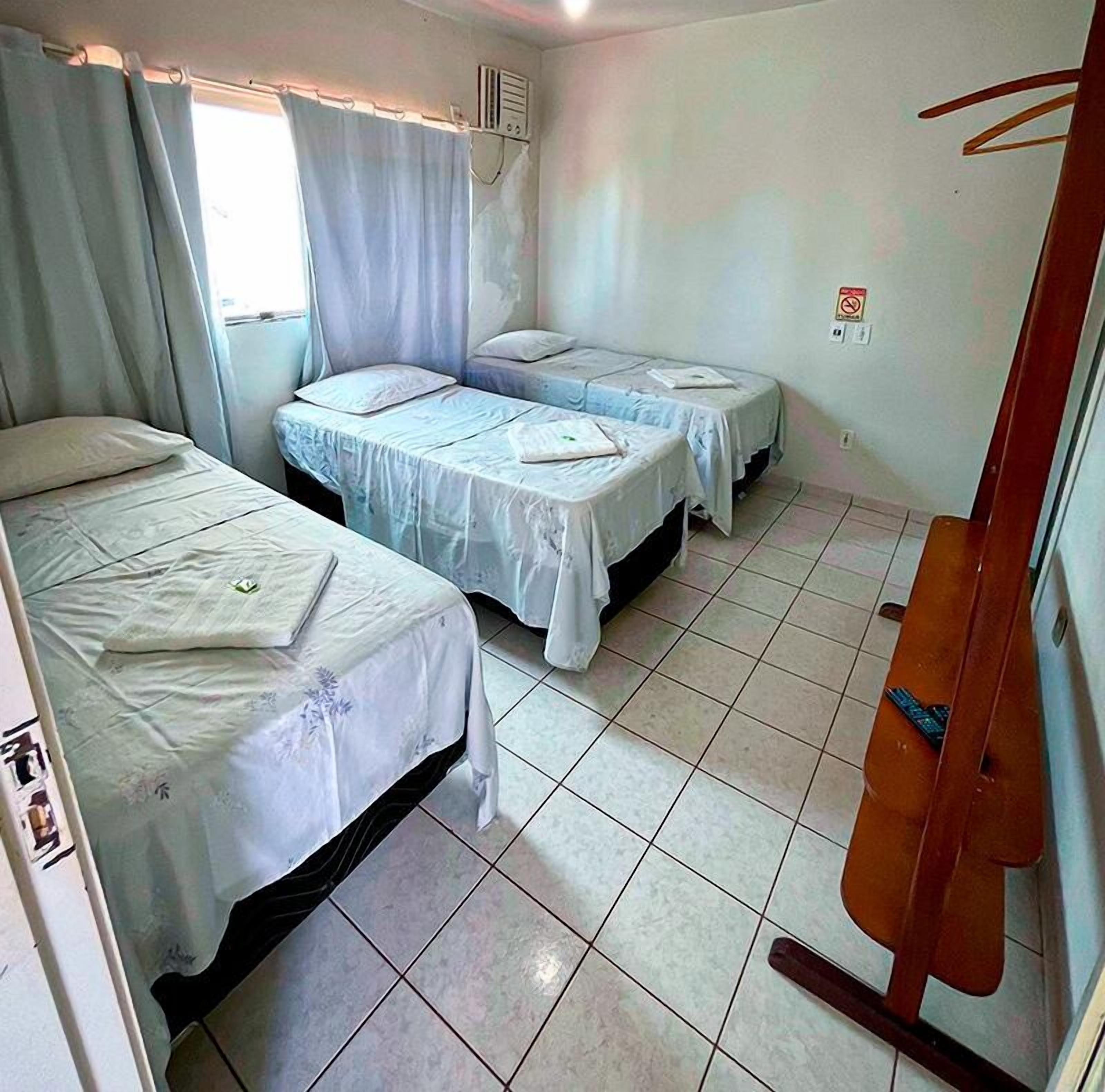 Una habitación pequeña y limpia con dos camas individuales, una ventana con cortinas, un radiador y piso de baldosas.