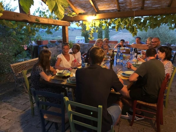 Un grupo de personas cenando en una mesa al aire libre, bajo una pérgola rodeada de vegetación.