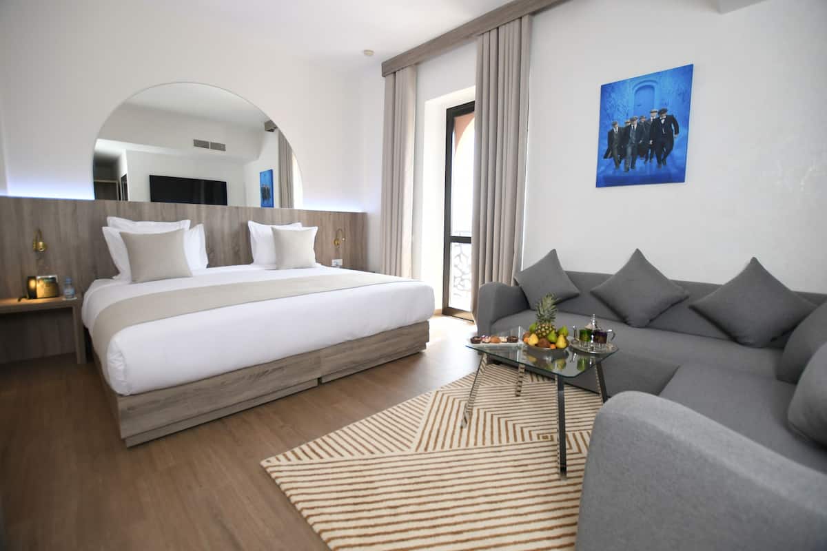 A modern hotel room with a large bed, a sofa, a glass coffee table, and a painting on the wall.