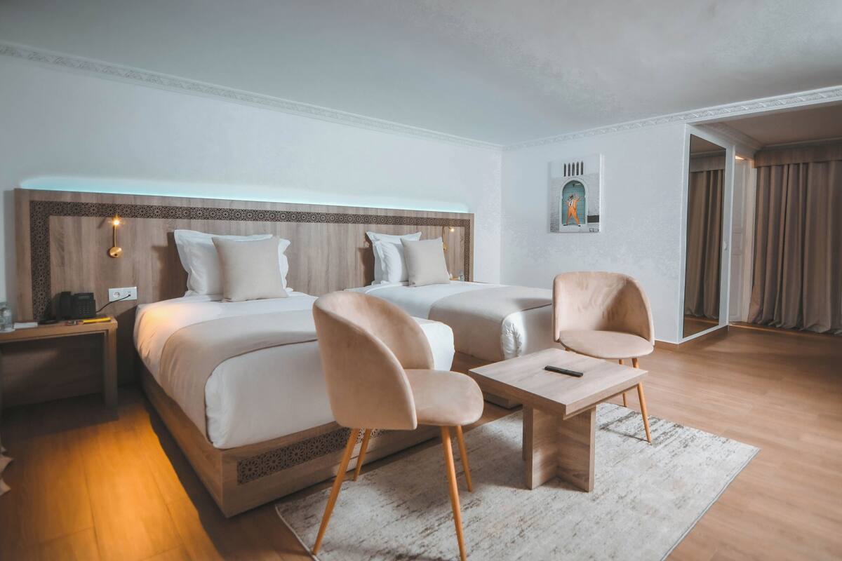 A hotel room with two beds, a small table, two chairs, a nightstand, and a wall-mounted lamp.