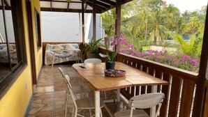 Een overdekt terras met een houten tafel en twee stoelen, een potplant en uitzicht op tropische begroeiing.