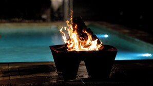 A poolside fire feature with flames, a pool, and a building in the background. - Casa D. Adelaide T1 (Azurara)