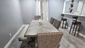 Un espace repas comprenant une longue table, des chaises en osier et un coin bar avec des tabourets.
