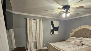 Une chambre à coucher avec un lit, un ventilateur de plafond, un téléviseur fixé au mur et une commode en bois.