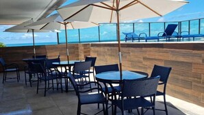 An outdoor seating area with tables and chairs, large umbrellas, and a view of the ocean. - Flat Nascente a passos do Mar (Ipojuca)