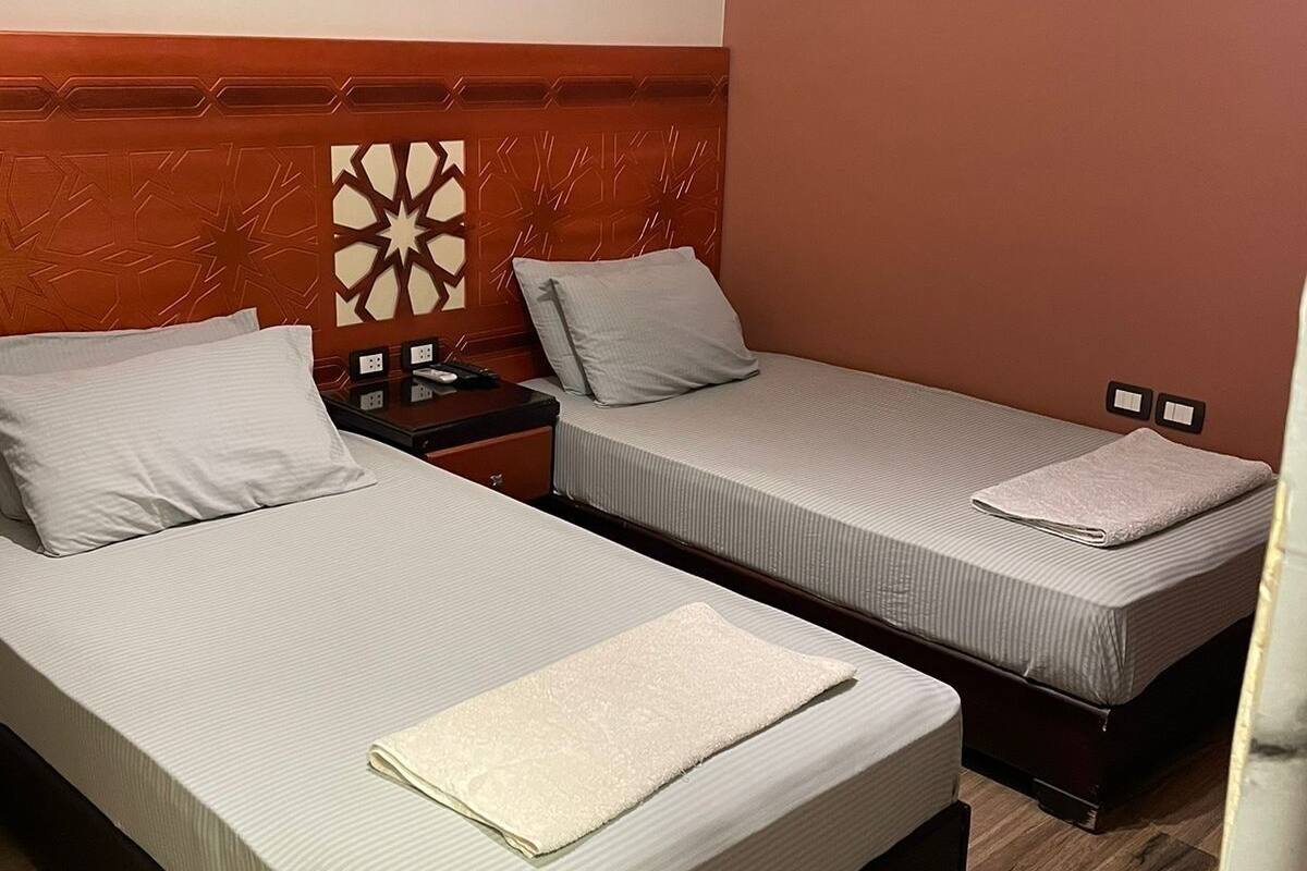 A hotel room with two single beds, a wooden headboard, a nightstand with a lamp, and a wall-mounted light fixture.