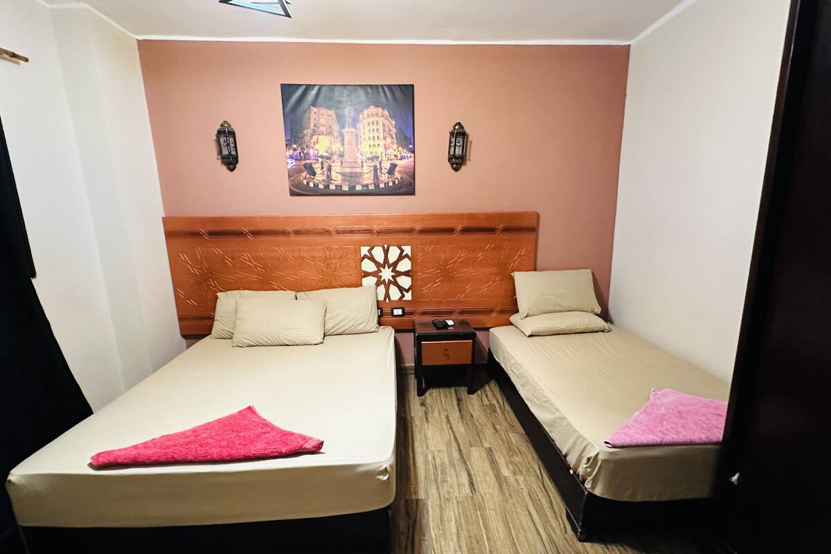 A hotel room with two beds, a nightstand, a wall-mounted lamp, and a painting of a cityscape.