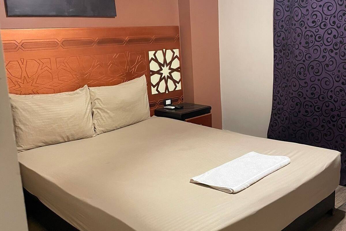 A neatly made bed with a white comforter and pillows, a headboard with a geometric pattern, a nightstand with a lamp, and a wall with a sunset skyline painting.