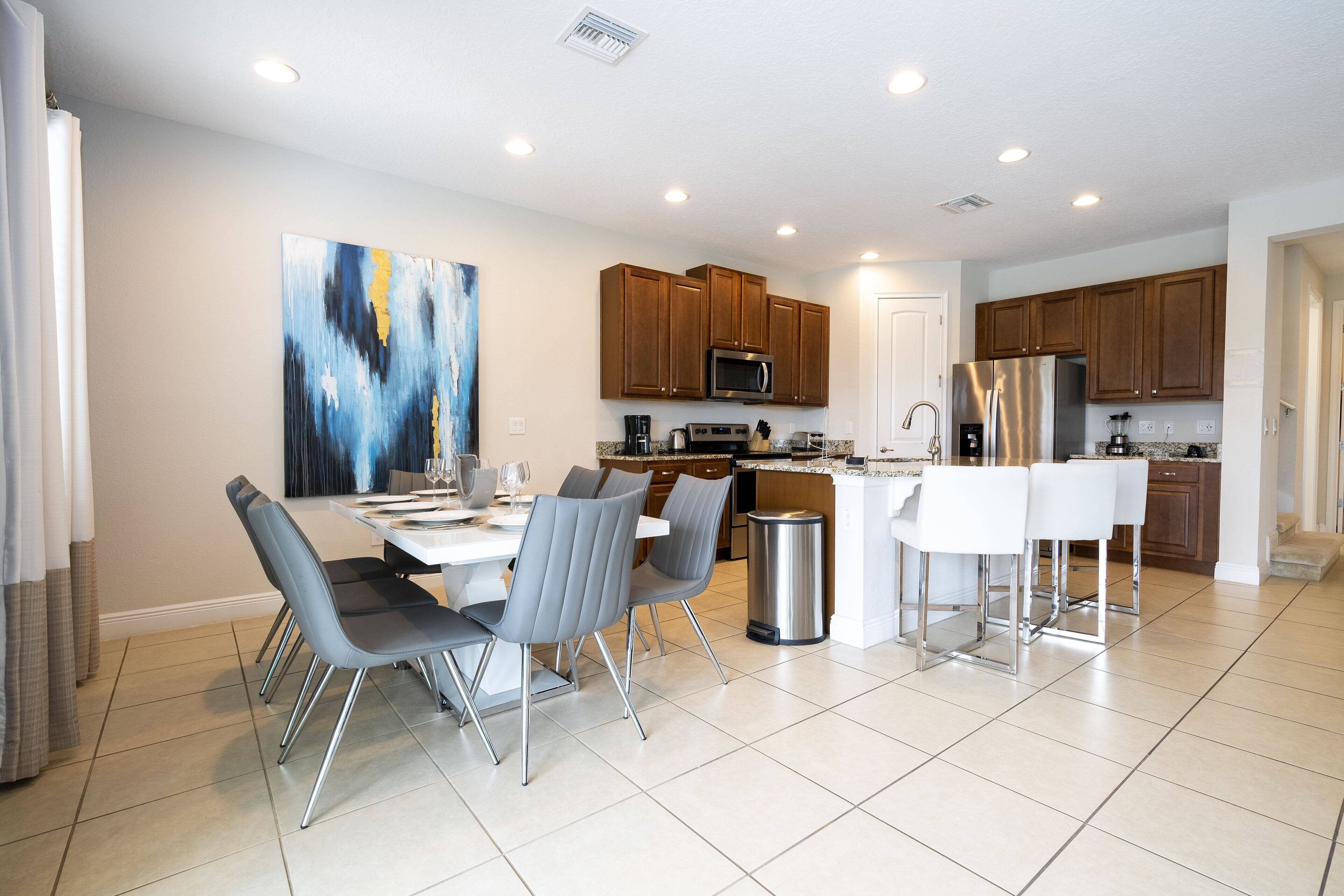 A modern kitchen with wooden cabinets, stainless steel appliances, and a dining area with grey chairs and a white table.