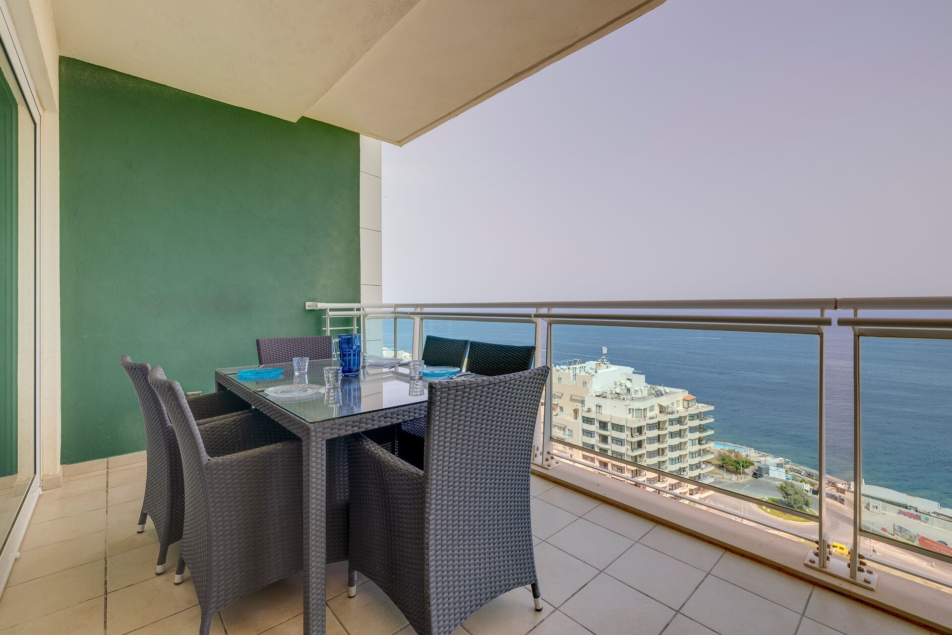 A balcony with a table and chairs, overlooking a coastal view with buildings and a clear sky.