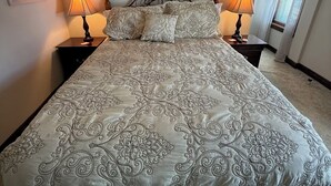 Una cama bien hecha con colcha estampada, dos lámparas de noche, un tapiz de macramé y un cuadro en la pared.