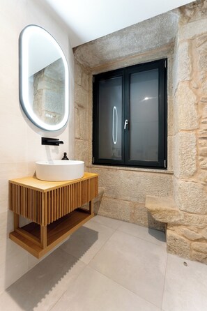 A bathroom with a stone wall, a wooden vanity with a white sink, a mirror, and a window with frosted glass doors. - Casa D. Adelaide T3 (Azurara)