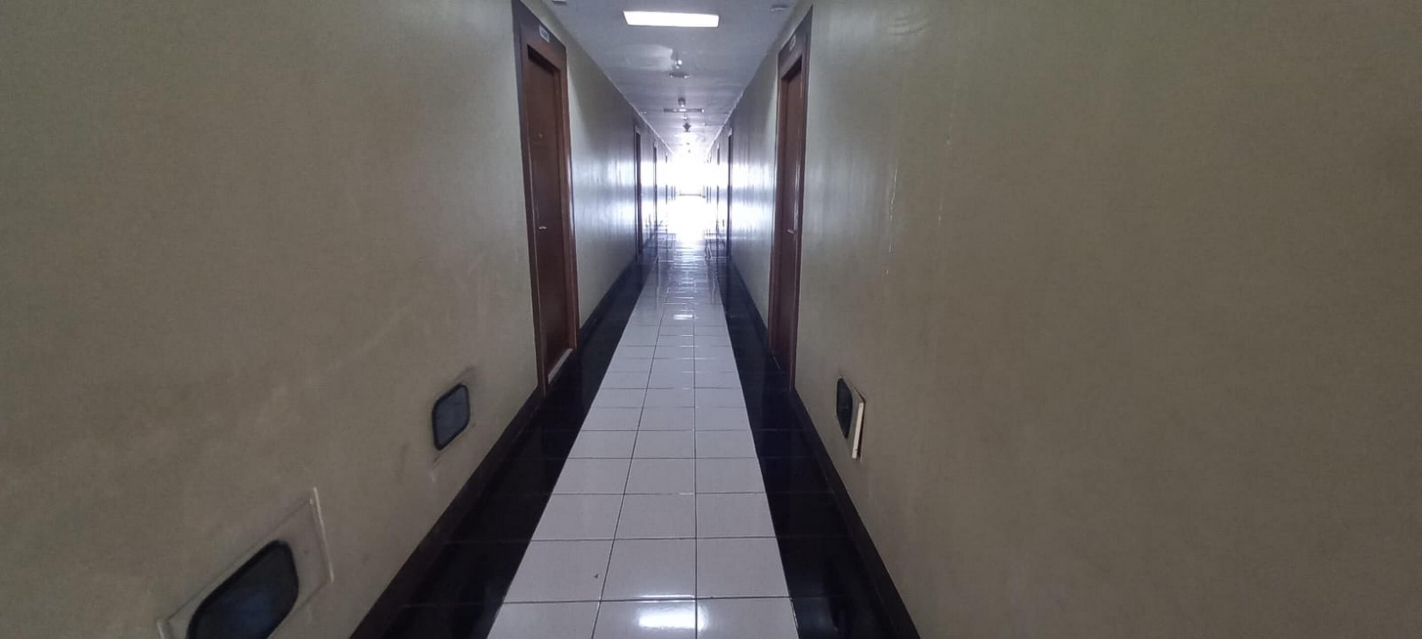 A long, narrow hallway with a tiled floor, flanked by beige walls and doors on both sides.