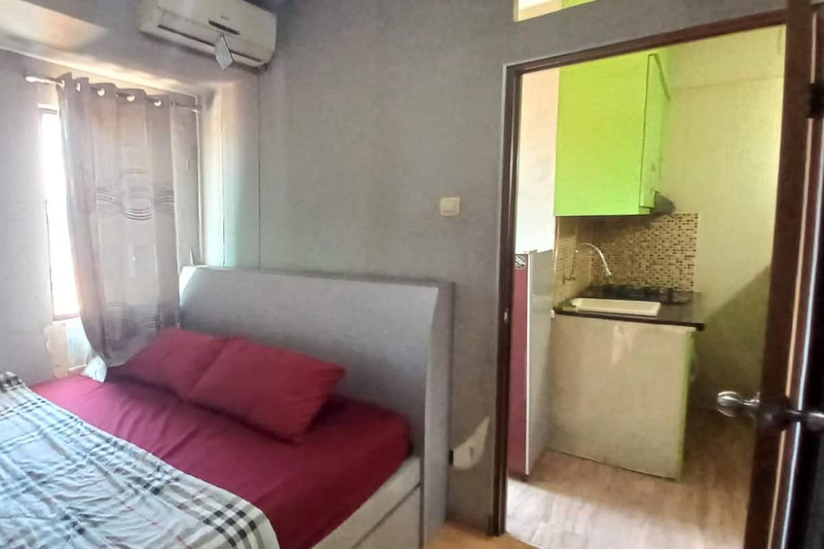 Um quarto com uma cama, uma janela com cortinas e uma cozinha compacta no cômodo vizinho.