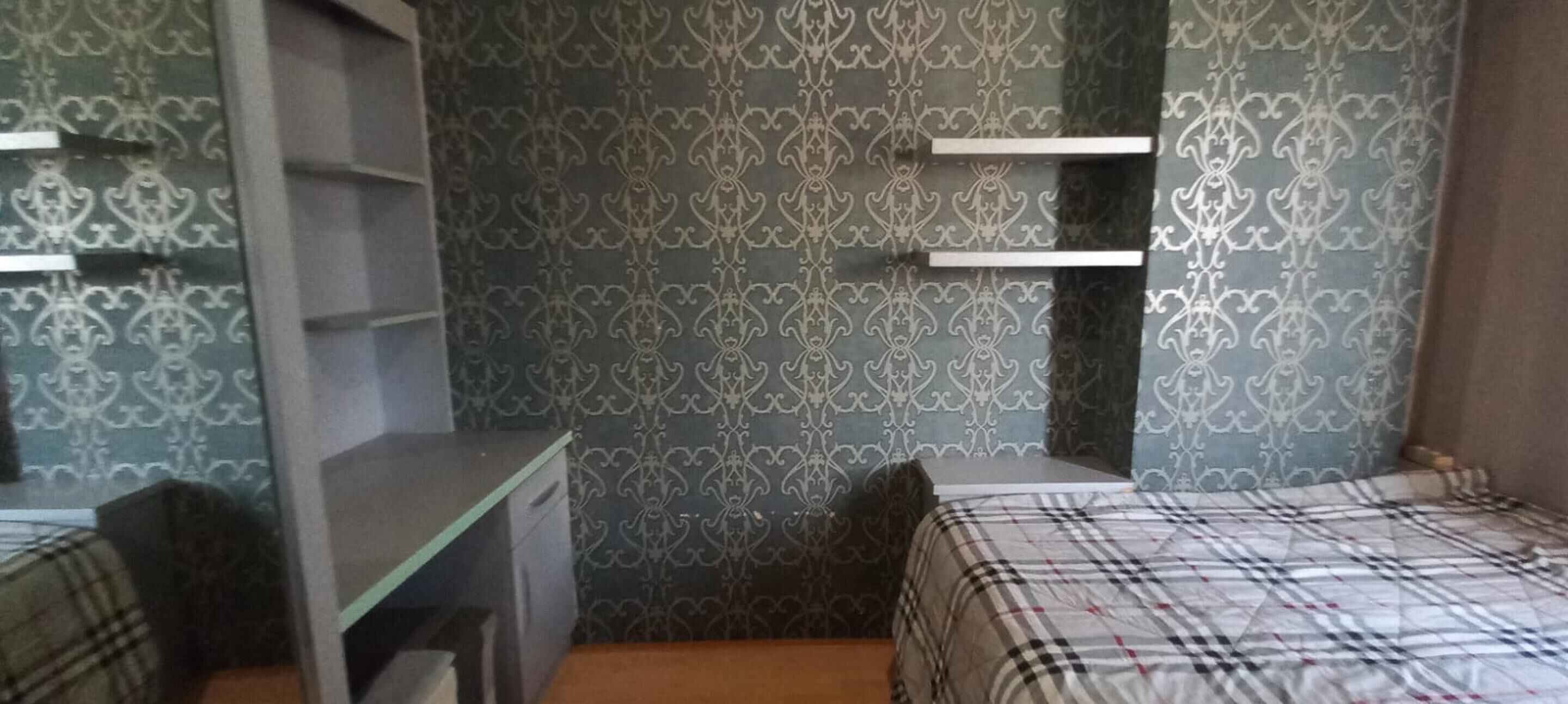 Ein Schlafzimmer mit einem Bett, einem Schreibtisch und einem Stuhl. An den Wänden befinden sich Regale und eine Wandtapete mit Muster.