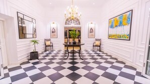 A hotel lobby with a checkered floor, a chandelier, framed artwork, and a central table with chairs.