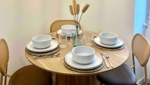 A round dining table with white plates, bowls, and cutlery, surrounded by wicker chairs. - Appartement T2 - Proche Centre et Lac (Aix-les-Bains)