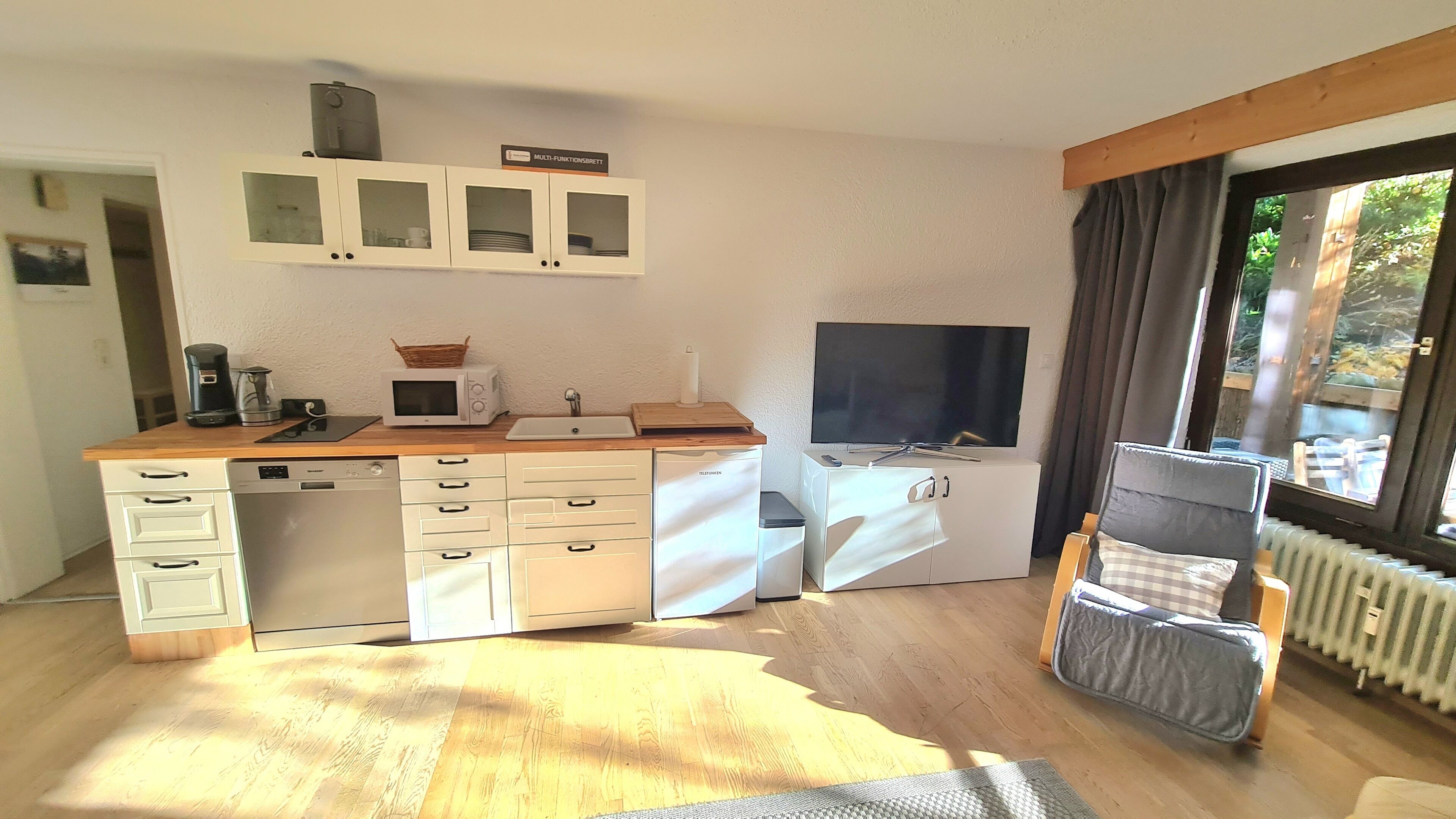 A compact kitchen with a microwave, coffee maker, and a small sink. There is a window with curtains, a radiator, and a TV mounted on the wall.