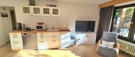 A compact kitchen with a microwave, coffee maker, and a small sink. There is a window with curtains, a radiator, and a TV mounted on the wall.