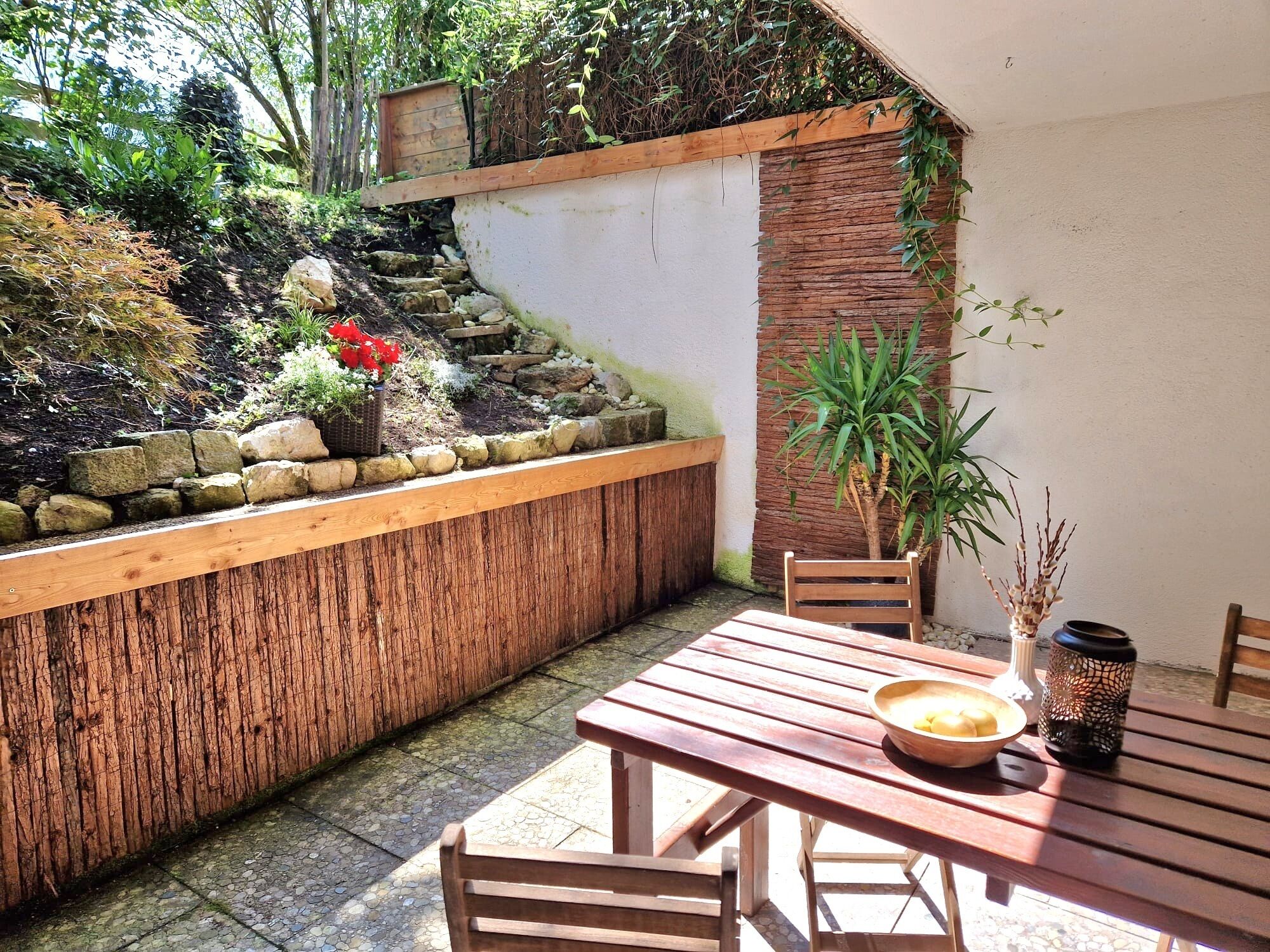 Une table et des chaises en bois sont disposées dans un coin du jardin, agrémenté d’un mur en pierre, de plantes et d’un petit banc en bois.