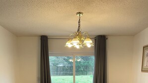 Een eetkamer met een houten tafel en stoelen, een kroonluchter en uitzicht op een gazon door grote ramen.
