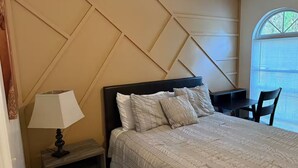 Een net opgemaakt bed met hoofdeinde, een nachtkastje met een lamp en een stoel bij een raam.