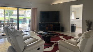 A living room with a TV, white leather sofas, a coffee table, and a ceiling fan. - CASA BELLA - HEATED POOL- HOT TUB- POOL TABLE -BBQ-PEACEFUL OASIS- 4 BEDS-  (Boynton Beach)