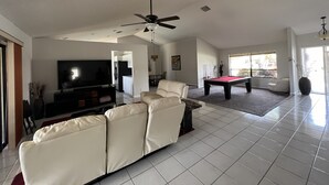 A spacious living room with a large flat-screen TV, a beige sectional sofa, a pool table, and a ceiling fan. - CASA BELLA - HEATED POOL- HOT TUB- POOL TABLE -BBQ-PEACEFUL OASIS- 4 BEDS-  (Boynton Beach)