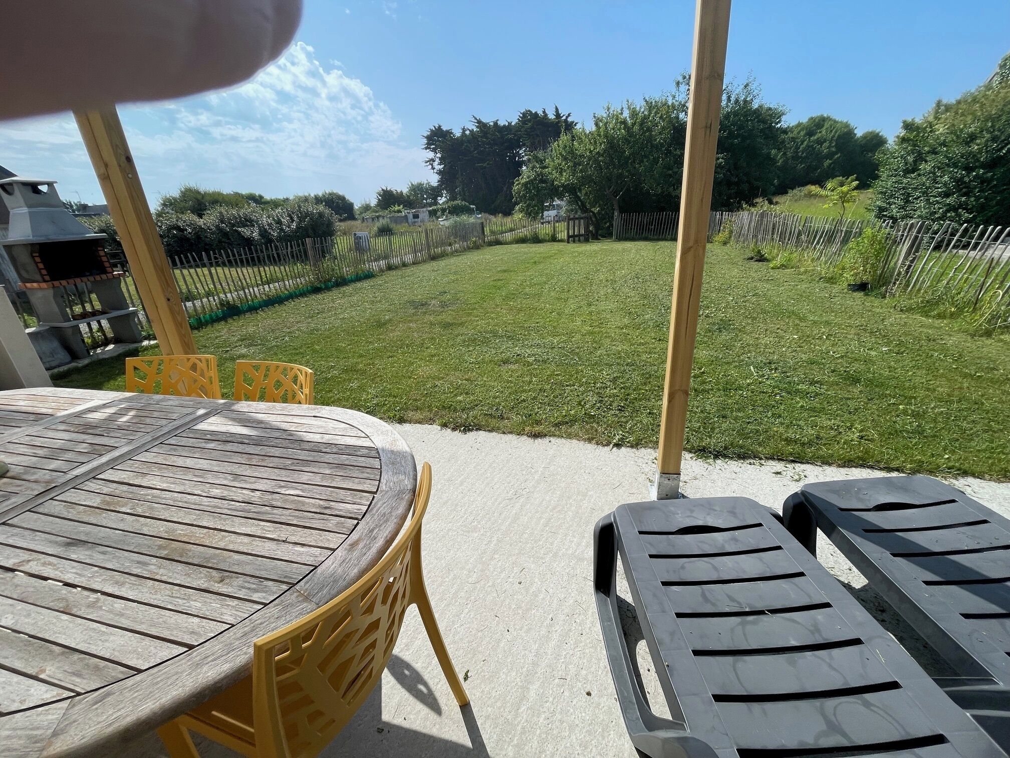A patio area with a round wooden table and yellow chairs, overlooking a grassy yard with a fence and trees.