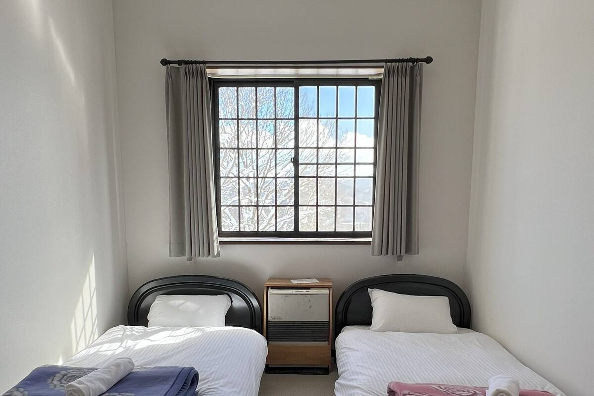 A room with two single beds, a window with curtains, a bedside table, and a radiator.