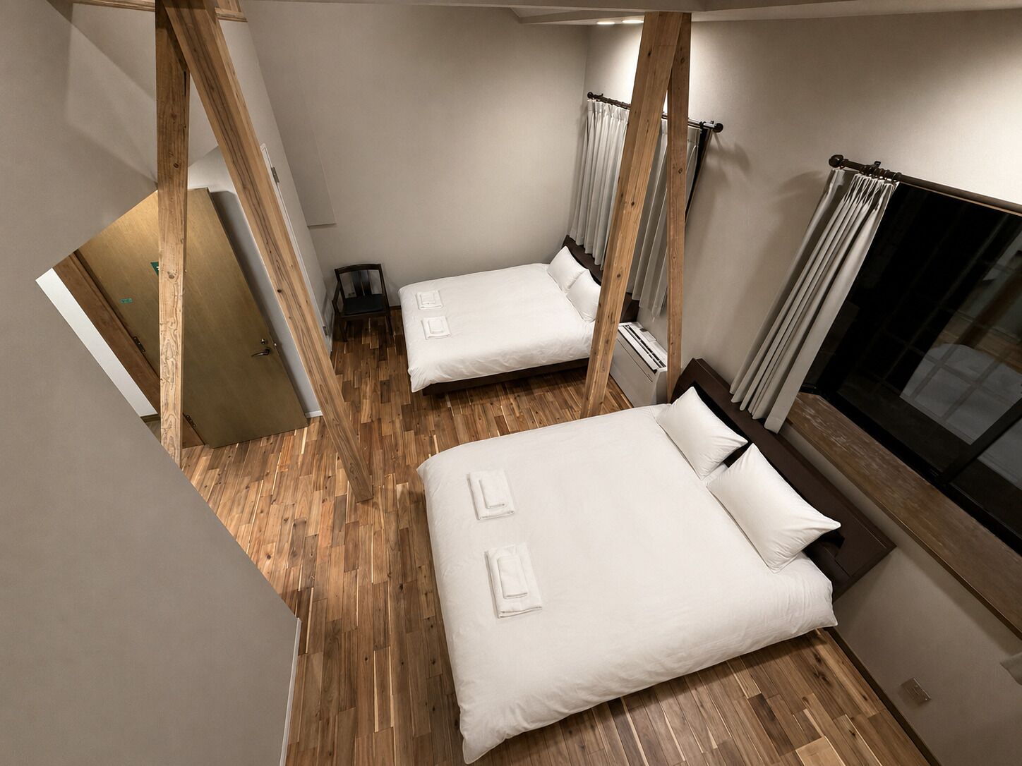 A bedroom with two beds, wooden flooring, a window with curtains, and a door leading to another room.