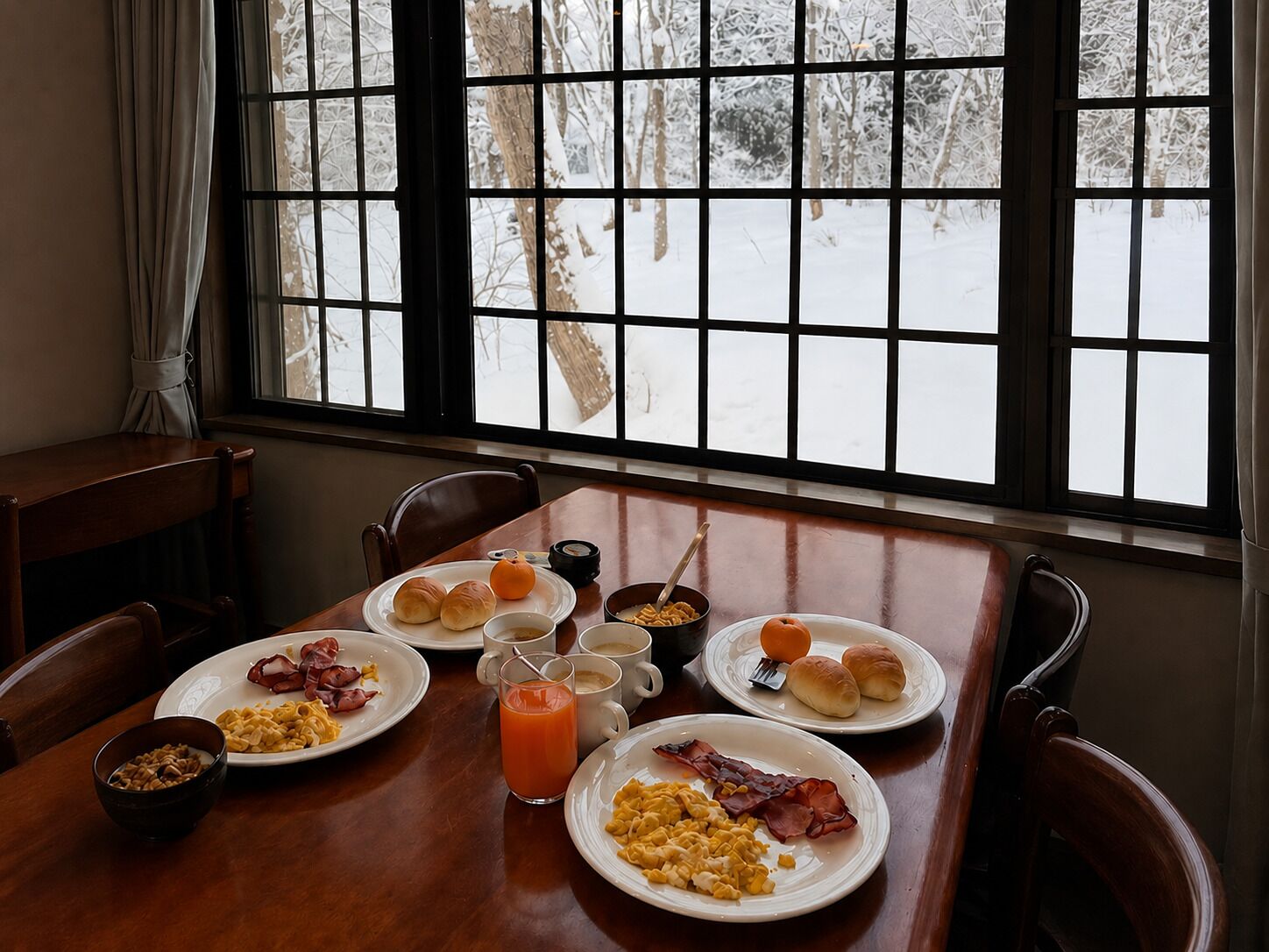 A dining table with breakfast plates, a glass of orange juice, and a view of a snowy landscape through a window.