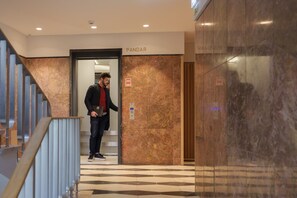 Um homem saindo de um elevador em um saguão moderno com paredes de mármore e um padrão geométrico no chão.