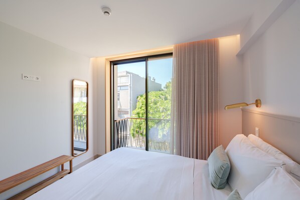 Una moderna camera d'hotel con un ampio letto, una panca, uno specchio e un balcone con vista su edifici e vegetazione.