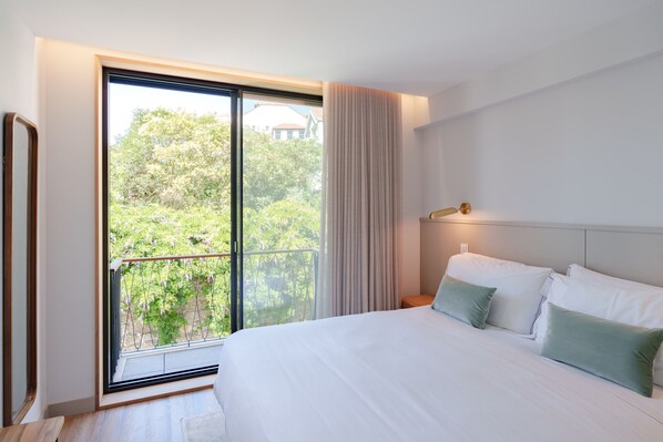 Habitación de hotel con una cama grande, una mesita de noche, una lámpara y un ventanal corredizo que da a un balcón con vistas a zonas verdes.