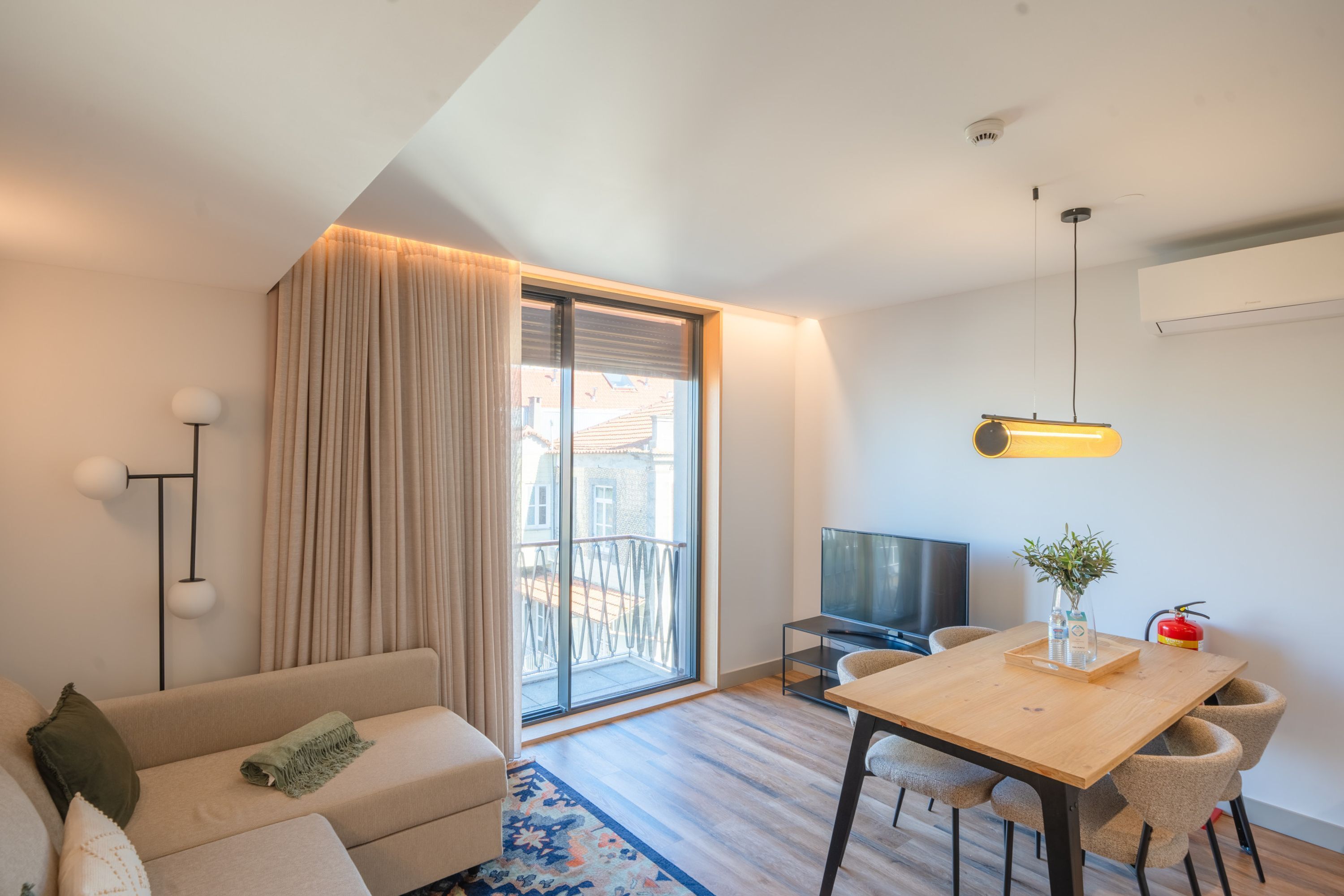 Una camera d'albergo moderna con divano, tavolo da pranzo, televisione e balcone con tende.