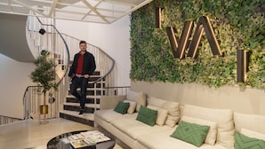 Un interior moderno con una escalera espiral, una pared verde con las letras iluminadas 'I.V.AI', un sofá beige con cojines verdes y una mesa de centro con revistas.