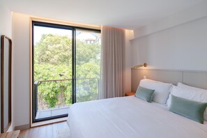 Une chambre d’hôtel moderne avec un grand lit, une table de chevet, une lampe et une vue sur la verdure grâce à une porte-fenêtre coulissante.