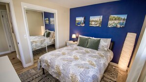 Un dormitorio con cama, mesita de noche, lámpara y espejo. Hay tres cuadros enmarcados en la pared.